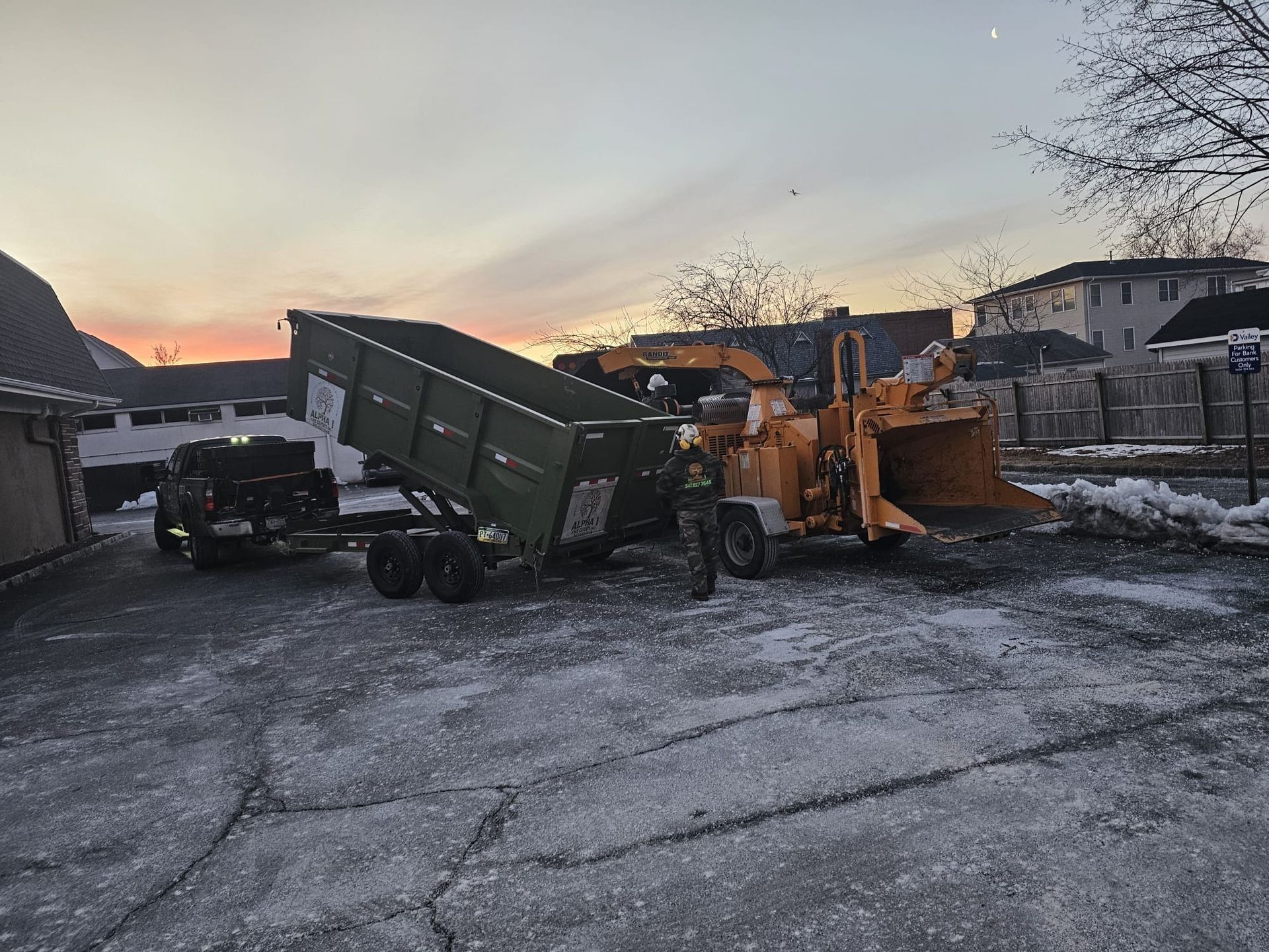 A wood chipper is loading wood chips into a green dump trailer in a snow-covered lot at dusk.