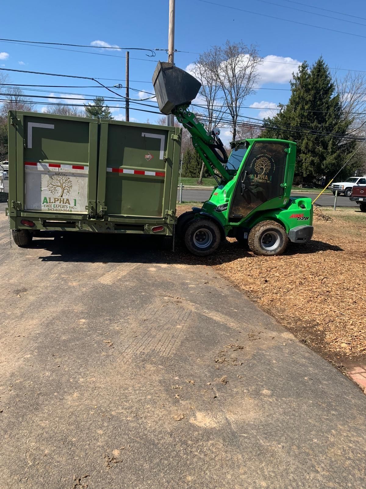 Green loader emptying contents into a dark green trailer on a sunny day. Asphalt and wood chips on the ground.