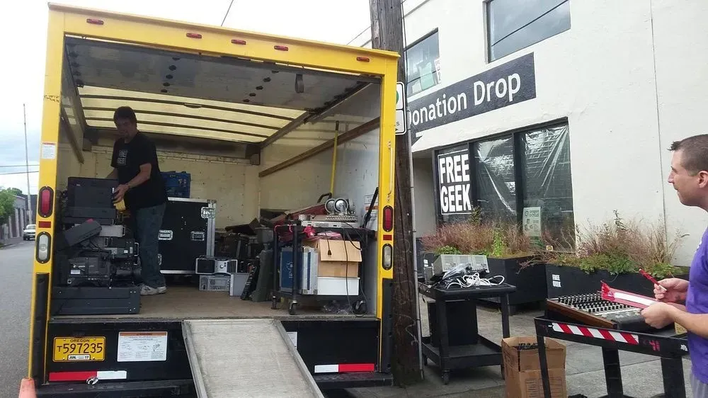 Yellow moving truck loading equipment outside a donation drop. People are loading items onto the truck, which is backed up to a building.