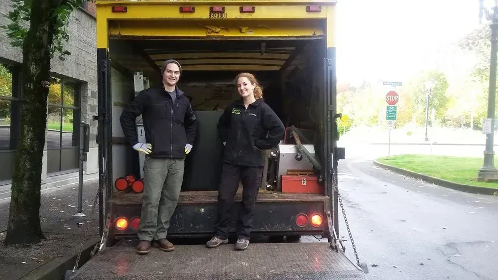 Two people standing in the open back of a yellow truck. The man and woman wear black jackets.