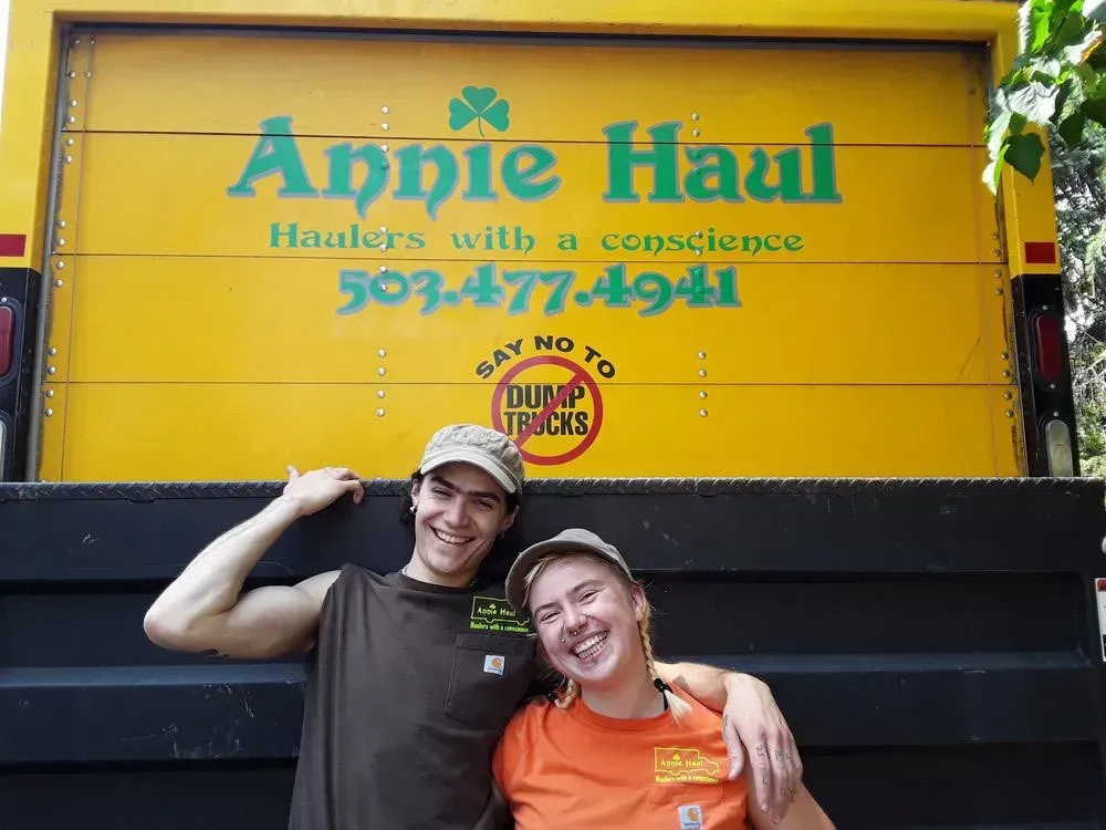 Two people smiling, posing in front of a yellow Annie Haul truck. The truck says
