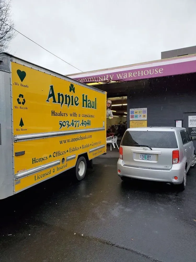 Yellow Annie Haul truck next to a silver minivan at a warehouse entrance on a rainy day.
