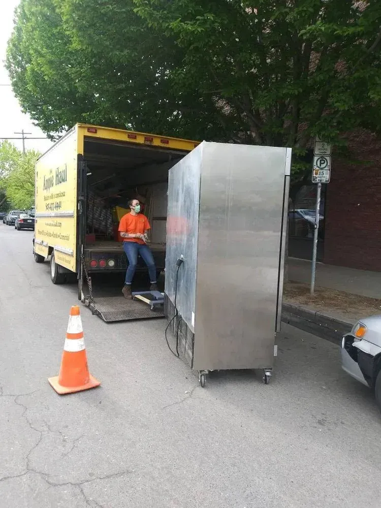 Truck unloading a large, stainless steel appliance; person in mask stands at the ramp, orange cone in street.