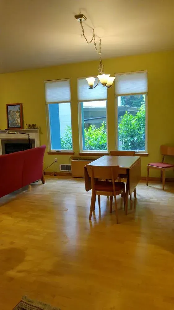 Living room with yellow walls, red sofa, dining table, and three windows overlooking greenery.