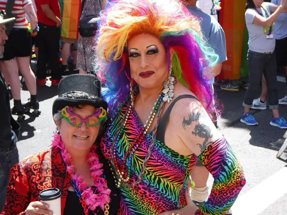 Two people at a Pride event: One in a rainbow dress and wig, the other with a hat and glasses, smiling.