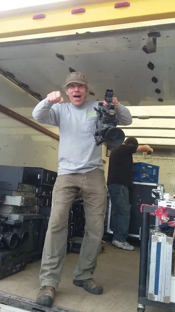 Man holding camera, standing in truck, excited expression. Another man in background. Equipment visible.