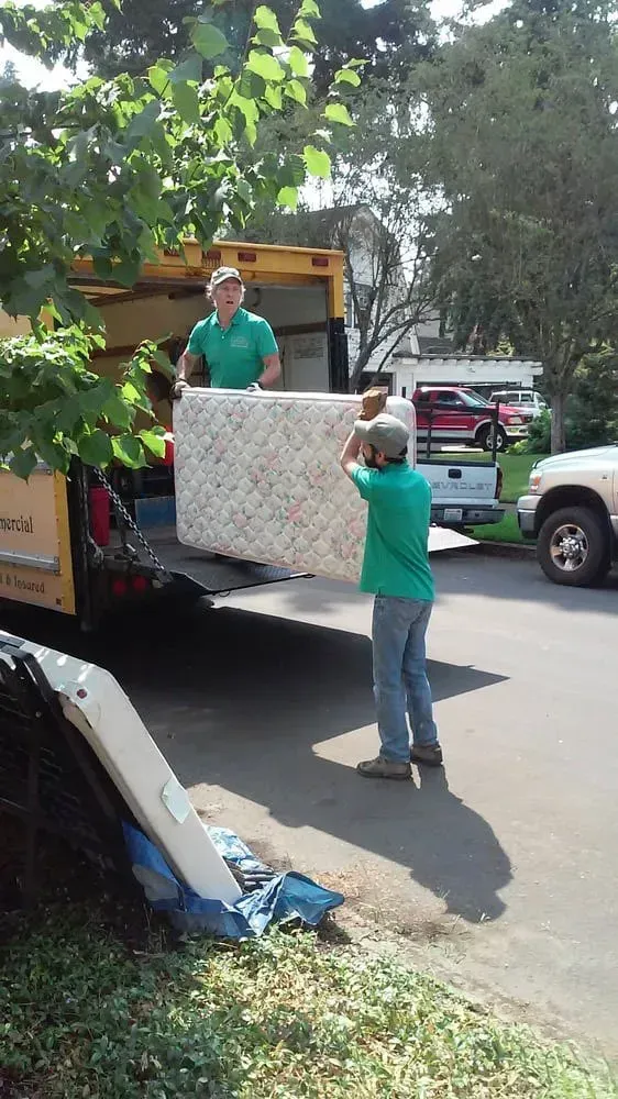 Two men in green shirts loading a mattress into a yellow truck on a sunny street.