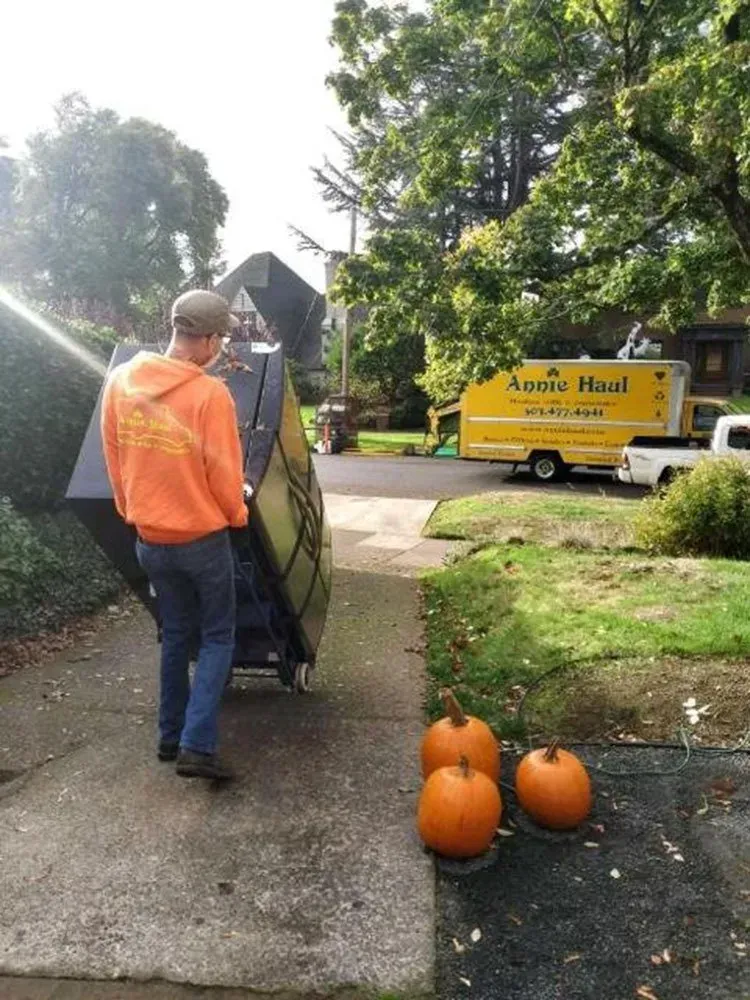 Man in orange hoodie rolls a piano on a dolly down a driveway, pumpkins sit in foreground, moving truck in the background.