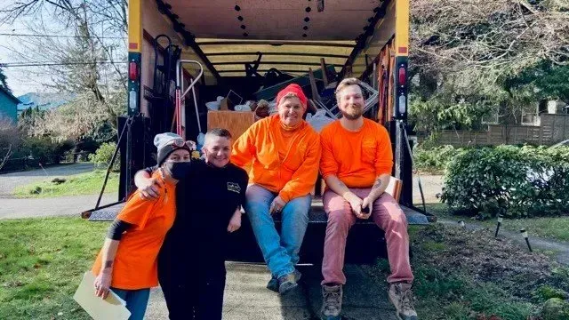 Four people pose in front of a yellow moving truck; three wear orange, one black. They are smiling and appear relaxed outdoors.
