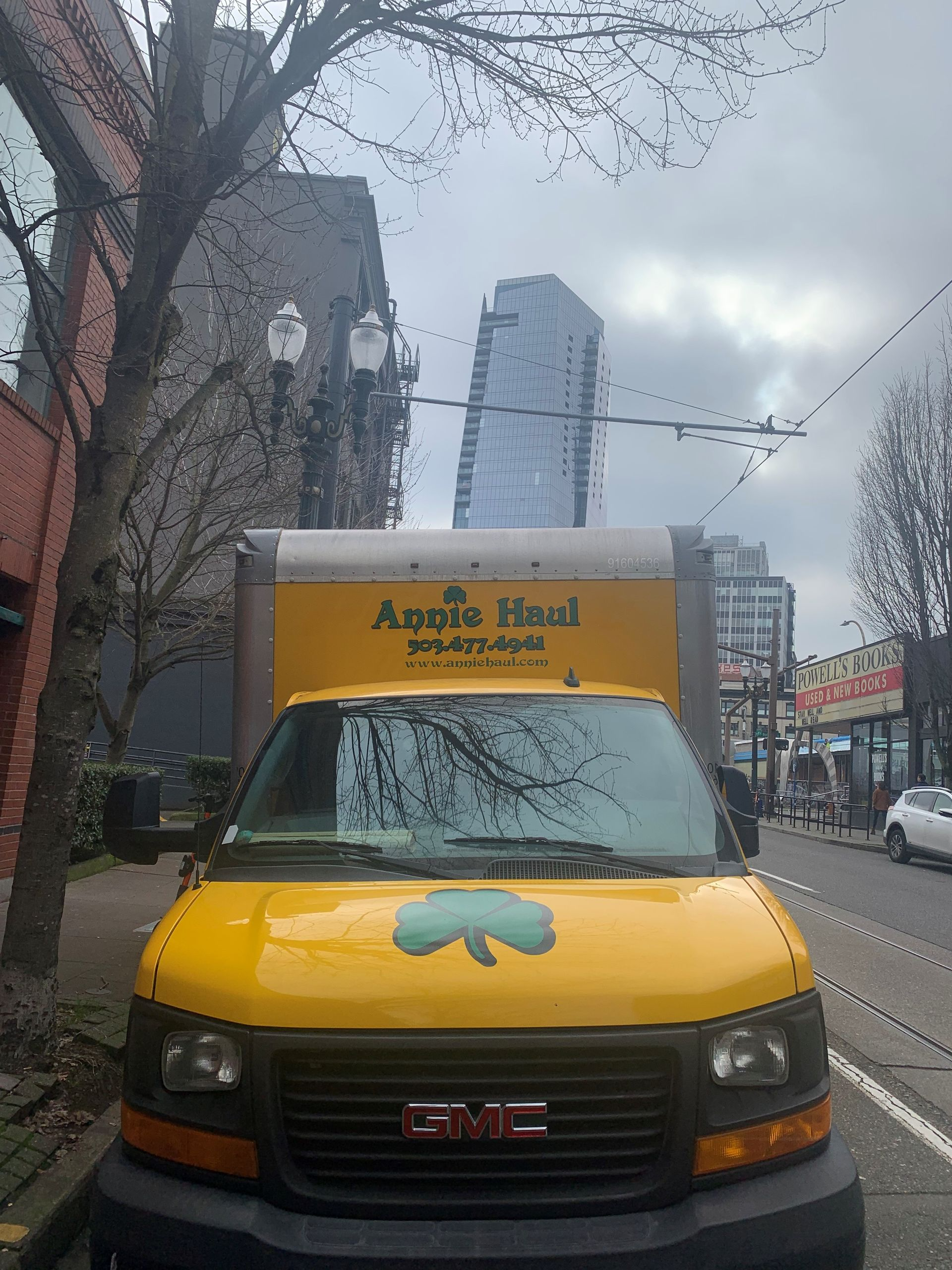 Yellow van with green shamrock parked on city street; tall buildings in background under cloudy sky.