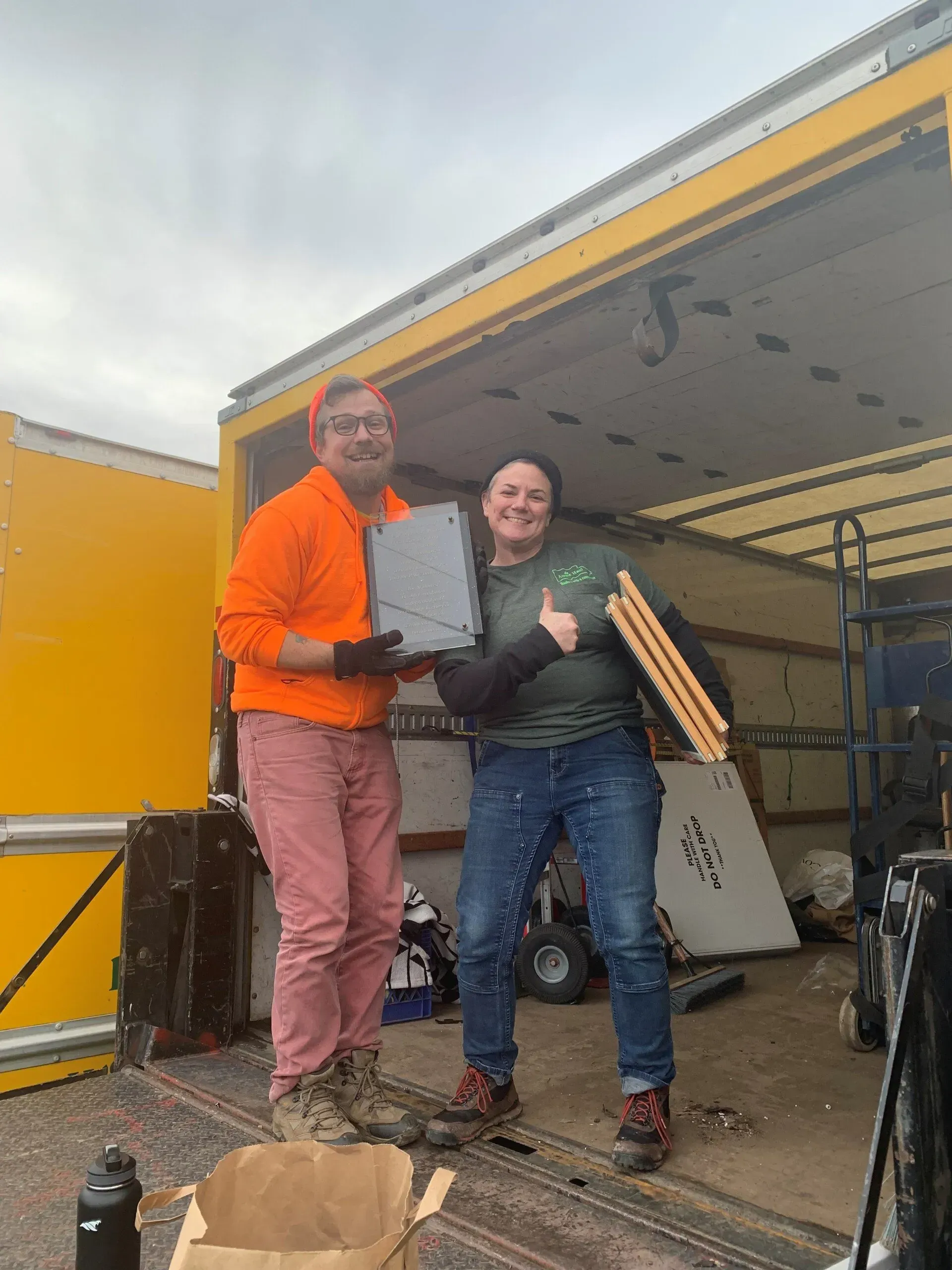 Two people stand inside a yellow truck, smiling while holding items. One in orange, one in green.