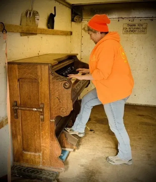 Woman playing an antique organ inside a weathered structure, wearing an orange hoodie and jeans.