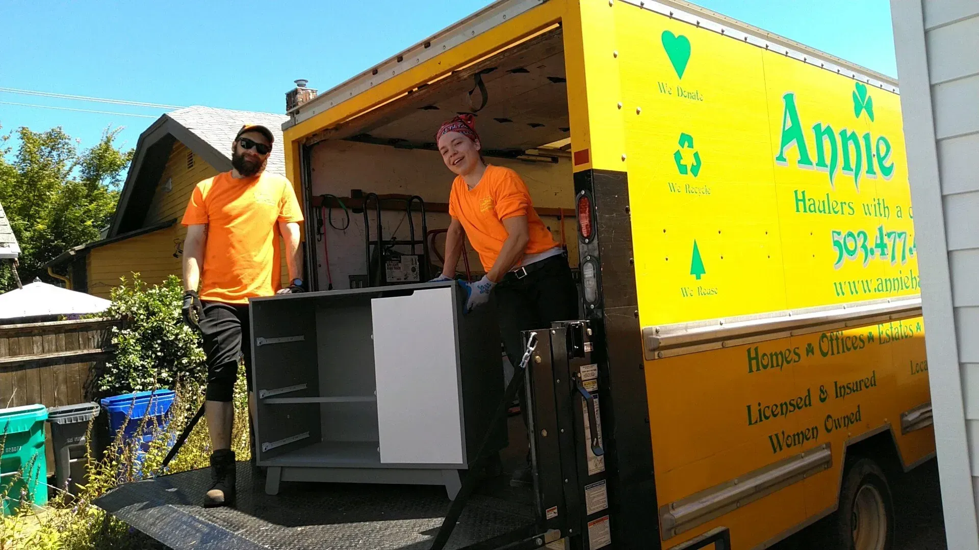 Two movers in orange shirts unload a cabinet from a yellow truck with 