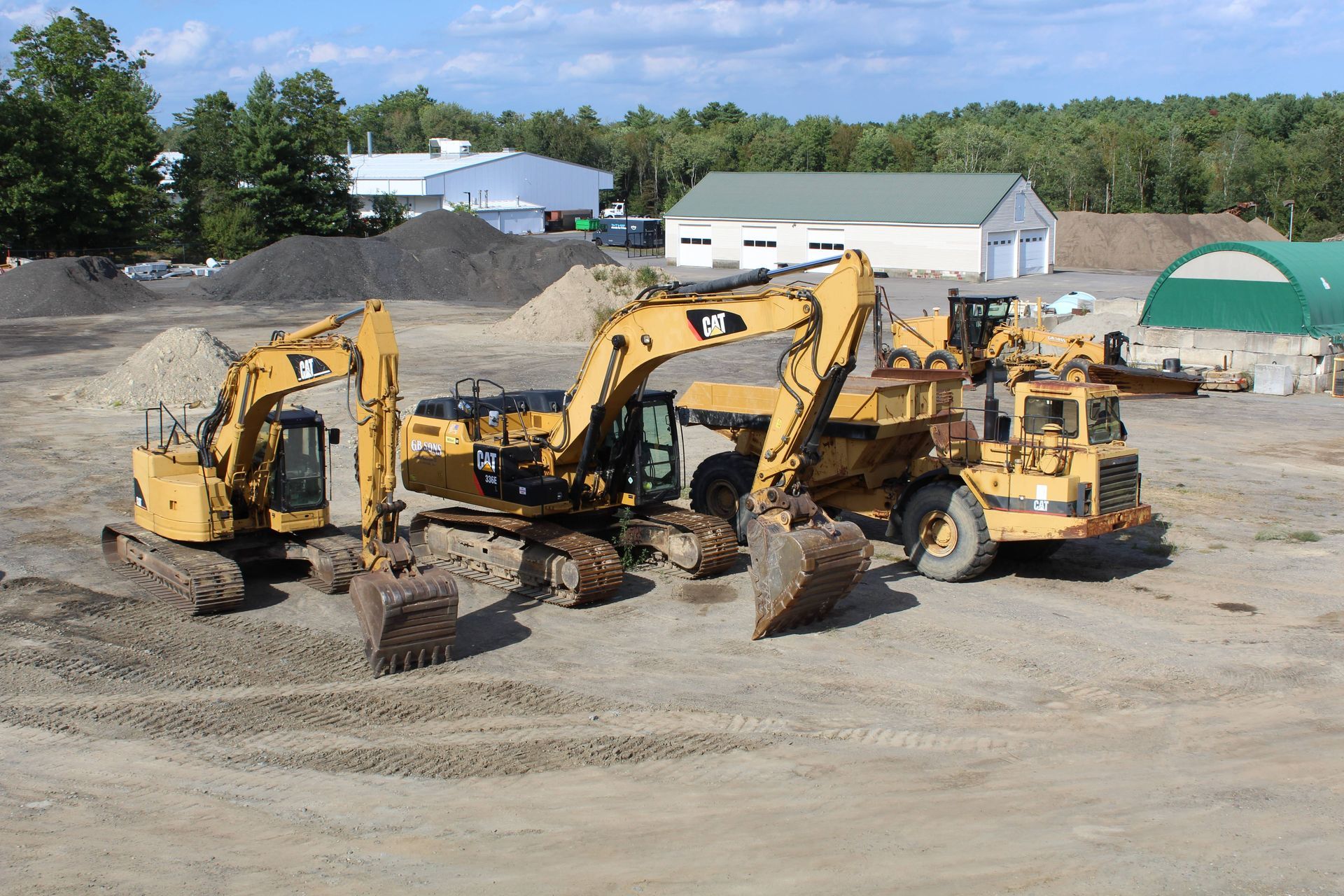 Yellow construction vehicles on a dirt lot, with piles of gravel and a few buildings in the background.
