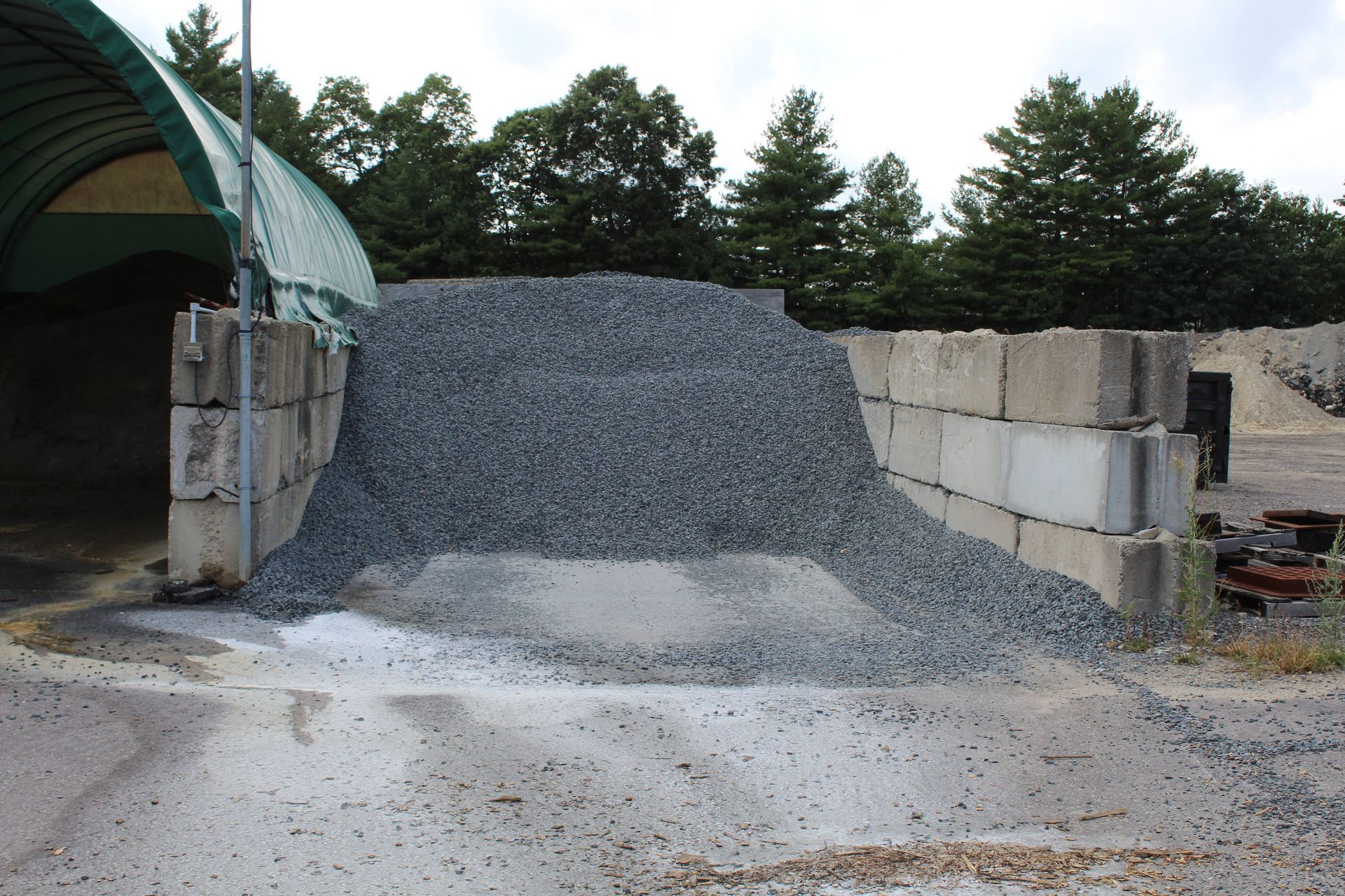 Pile of gray gravel next to a concrete block wall, under a green-roofed structure.