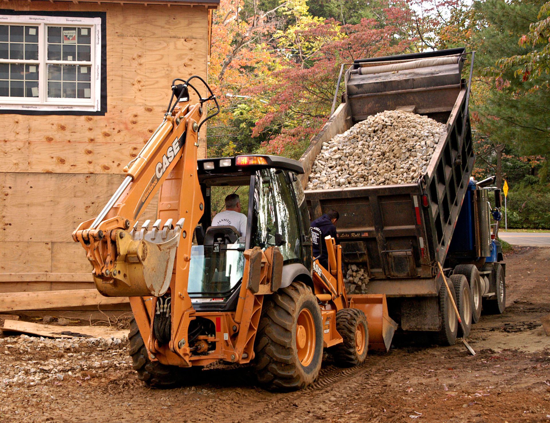 Orange backhoe loading gravel into a dump truck beside a building under construction.