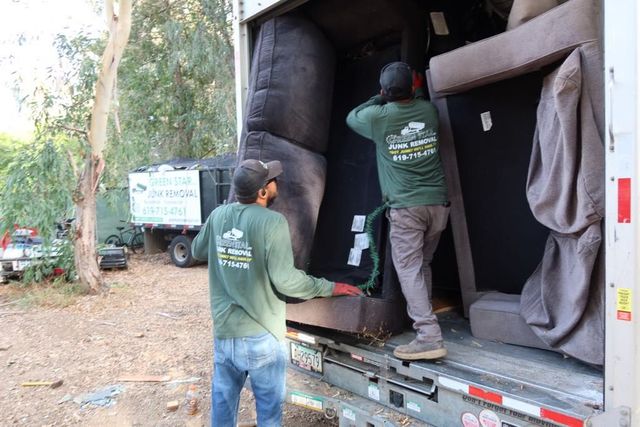 Two movers loading furniture into a truck. The men wear green shirts and are outdoors.