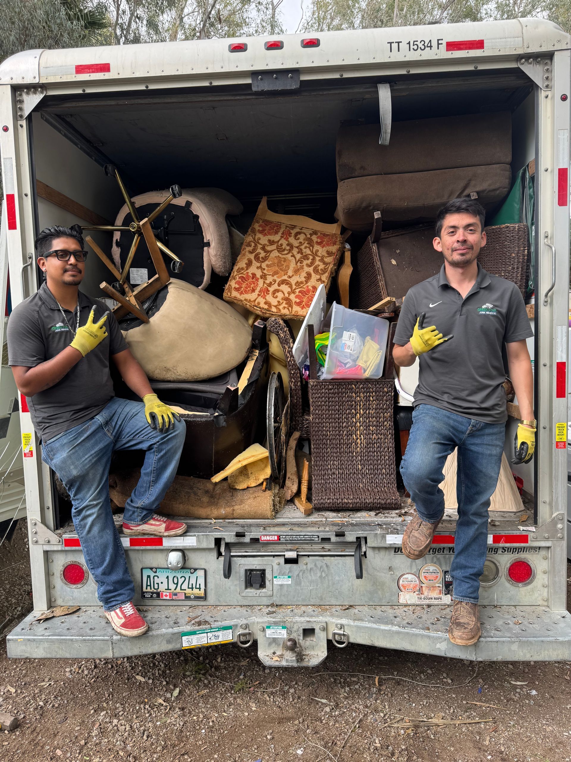 Two men in uniform pose with a loaded moving truck, holding up peace signs; truck bed is filled with furniture.