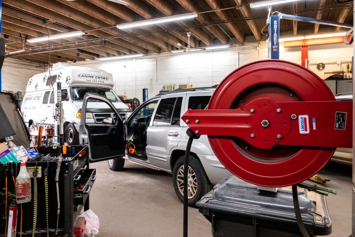 Car repair shop interior with a silver SUV and white van, red hose reel prominent in foreground.