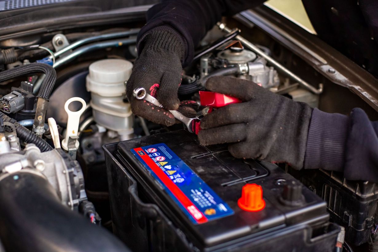 Hands in black gloves using wrenches to work on a car battery in an engine bay.