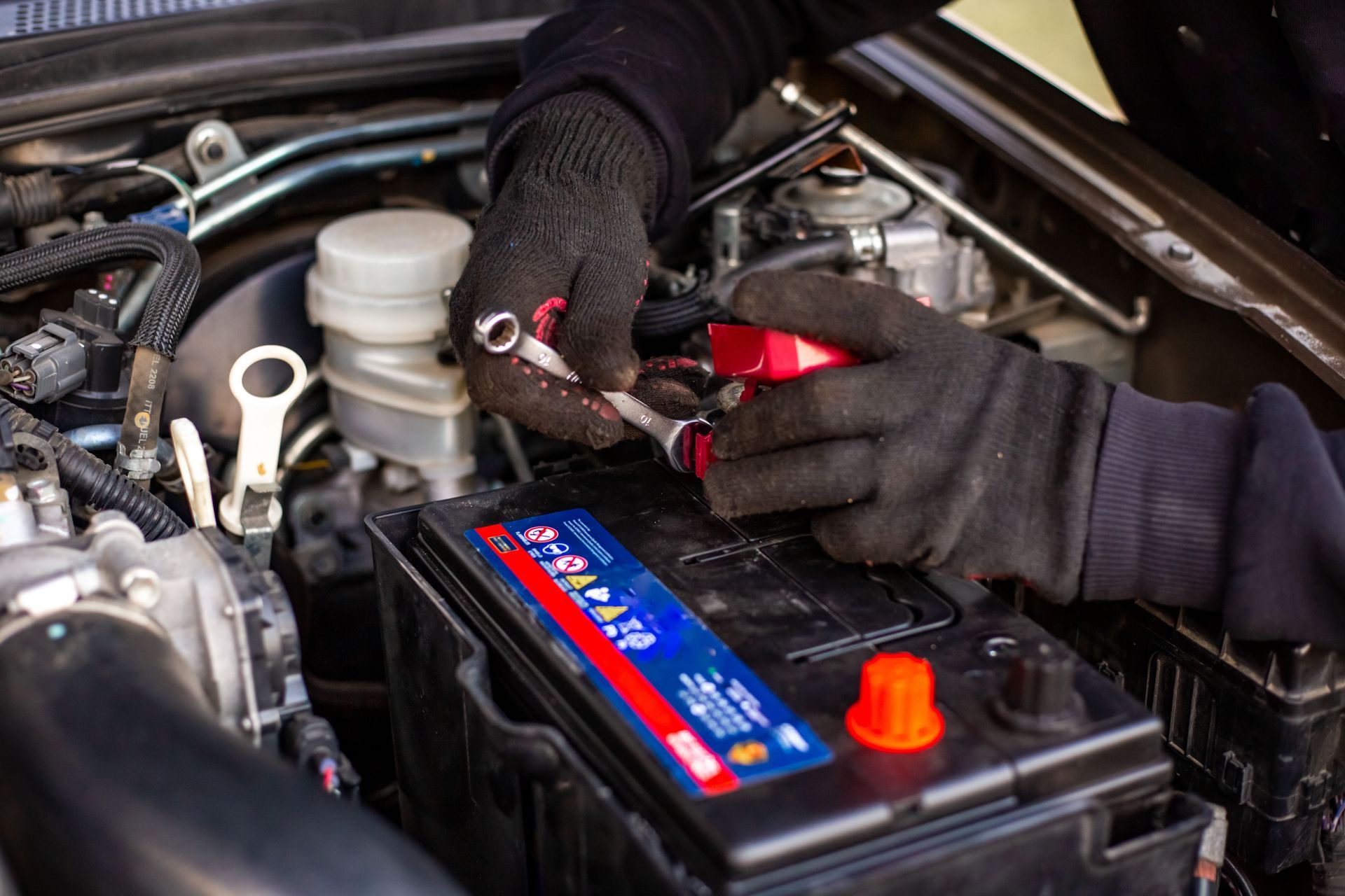 Hands in black gloves using wrenches to work on a car battery in an engine bay.