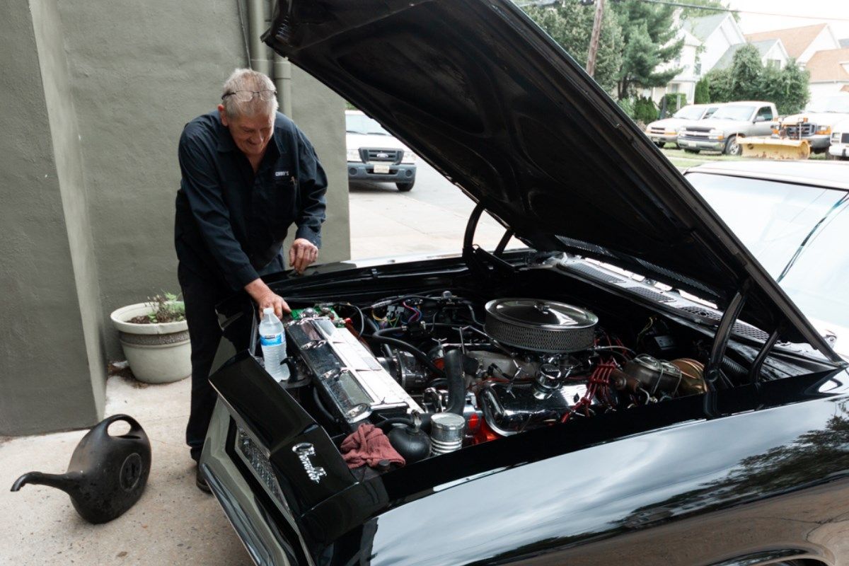 Man working on the engine of a black classic car outdoors, pouring fluid.