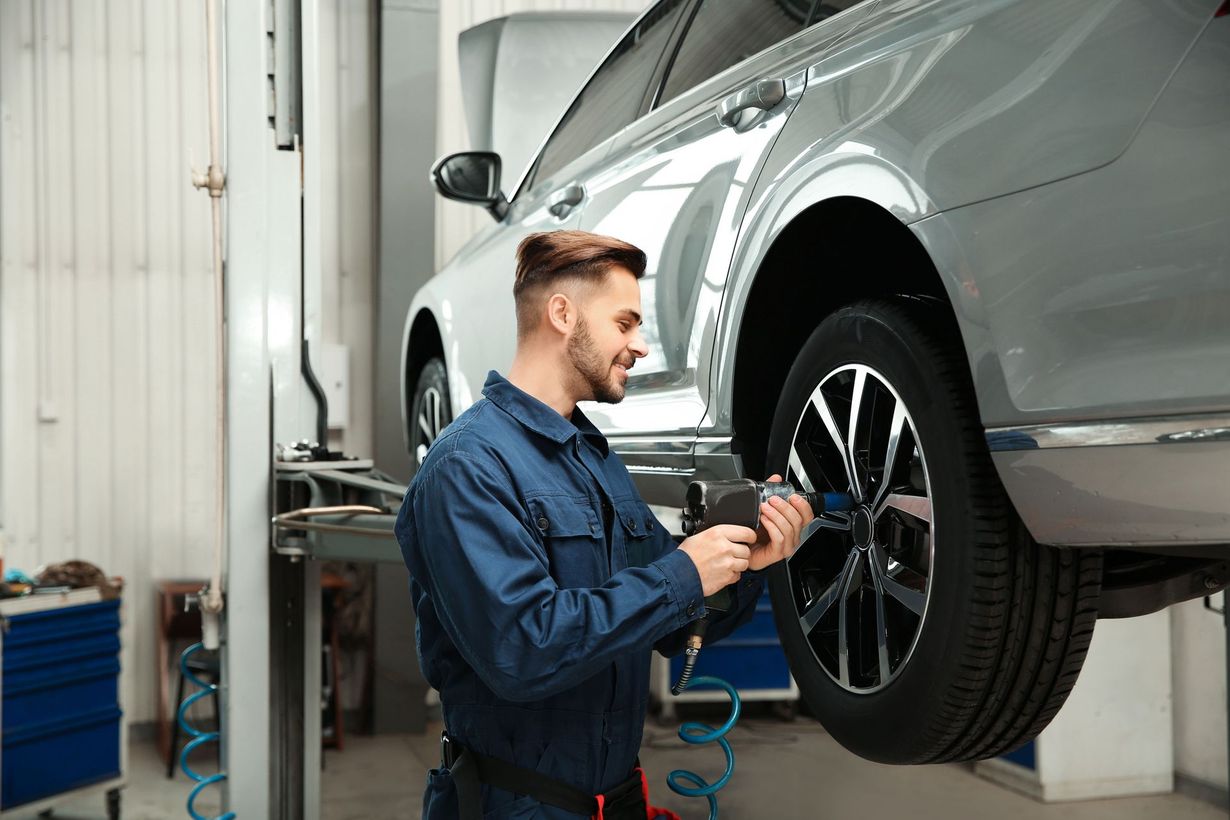 Mechanic in blue coveralls using a tool on a car tire in a garage.