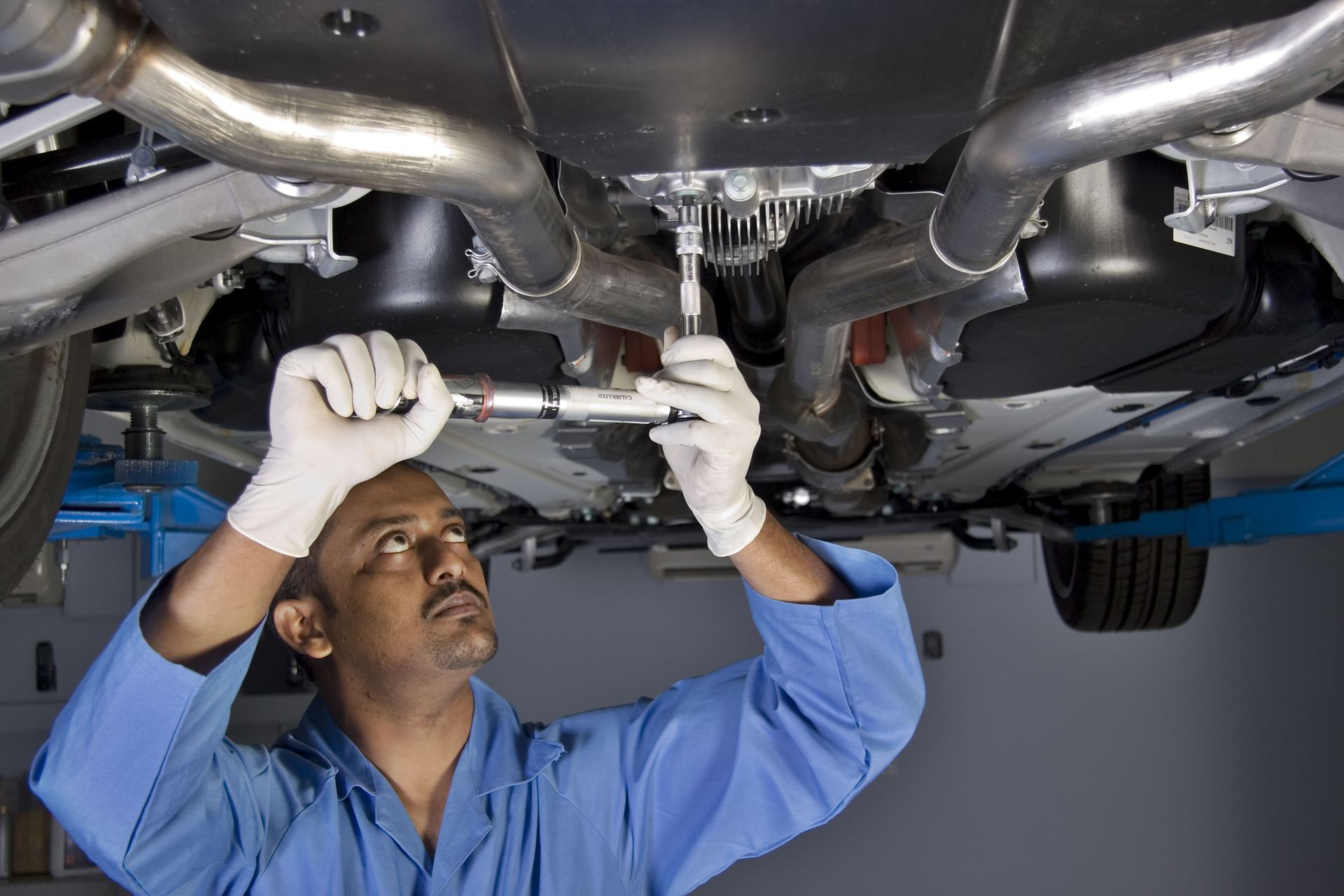 Mechanic in blue uniform works under car, using a wrench.