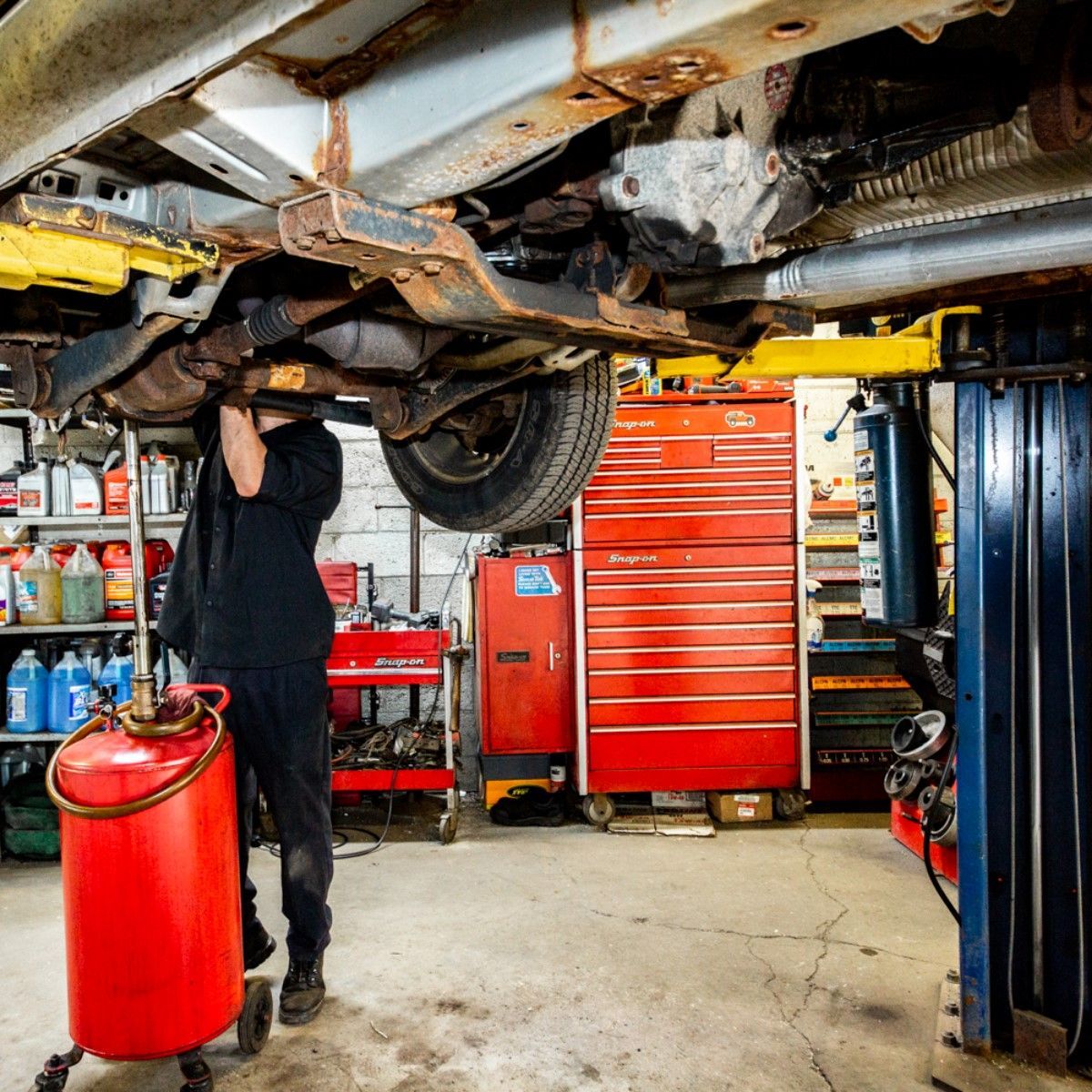 Mechanic working under a car on a lift in a garage, using a red fluid tank.