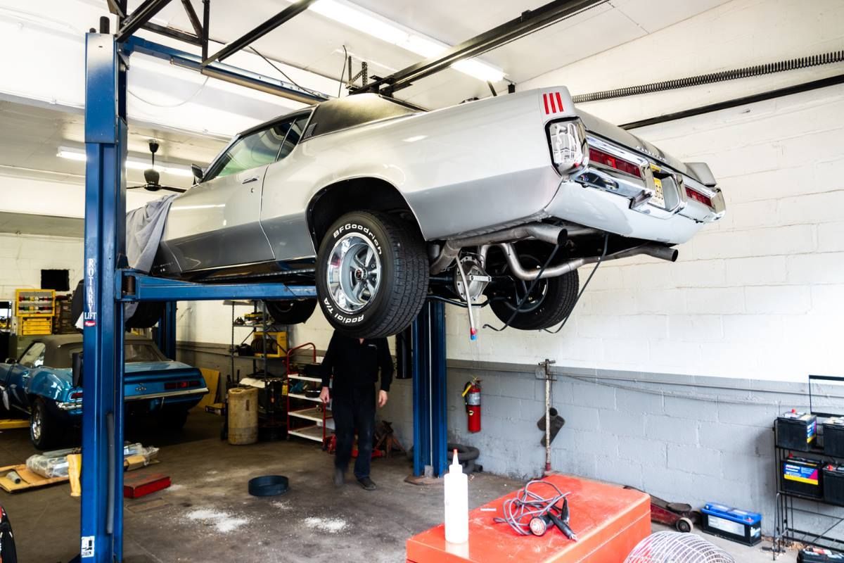 A classic silver car raised on a lift in a garage, a mechanic works underneath it.