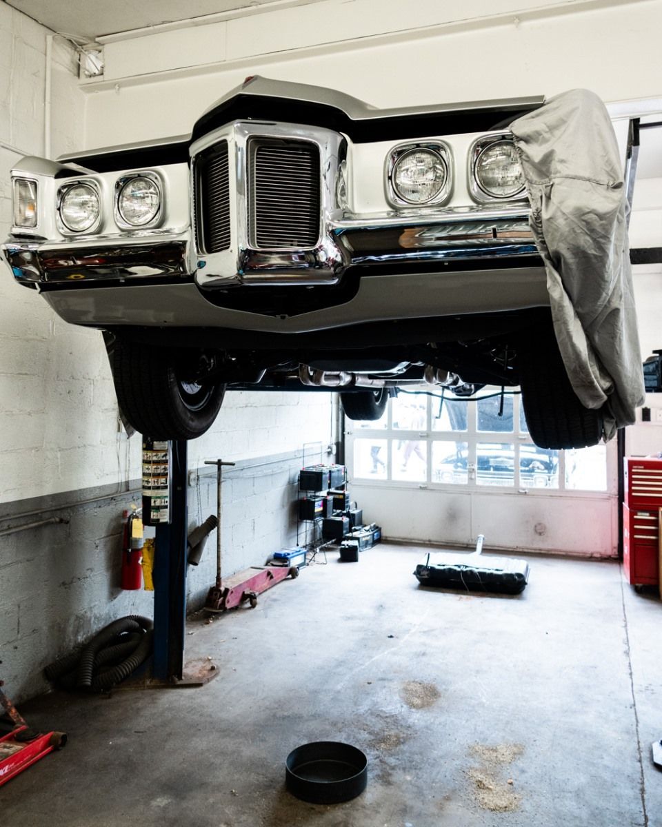 Classic car raised on a lift in a garage, lights on, covered front, tools visible.