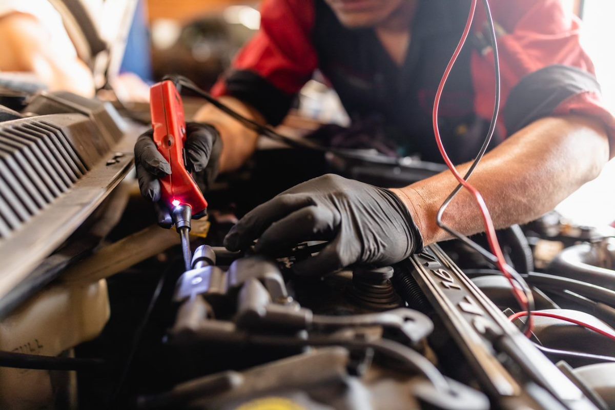 Mechanic in red jumpsuit using a probe on a car engine, wearing black gloves.