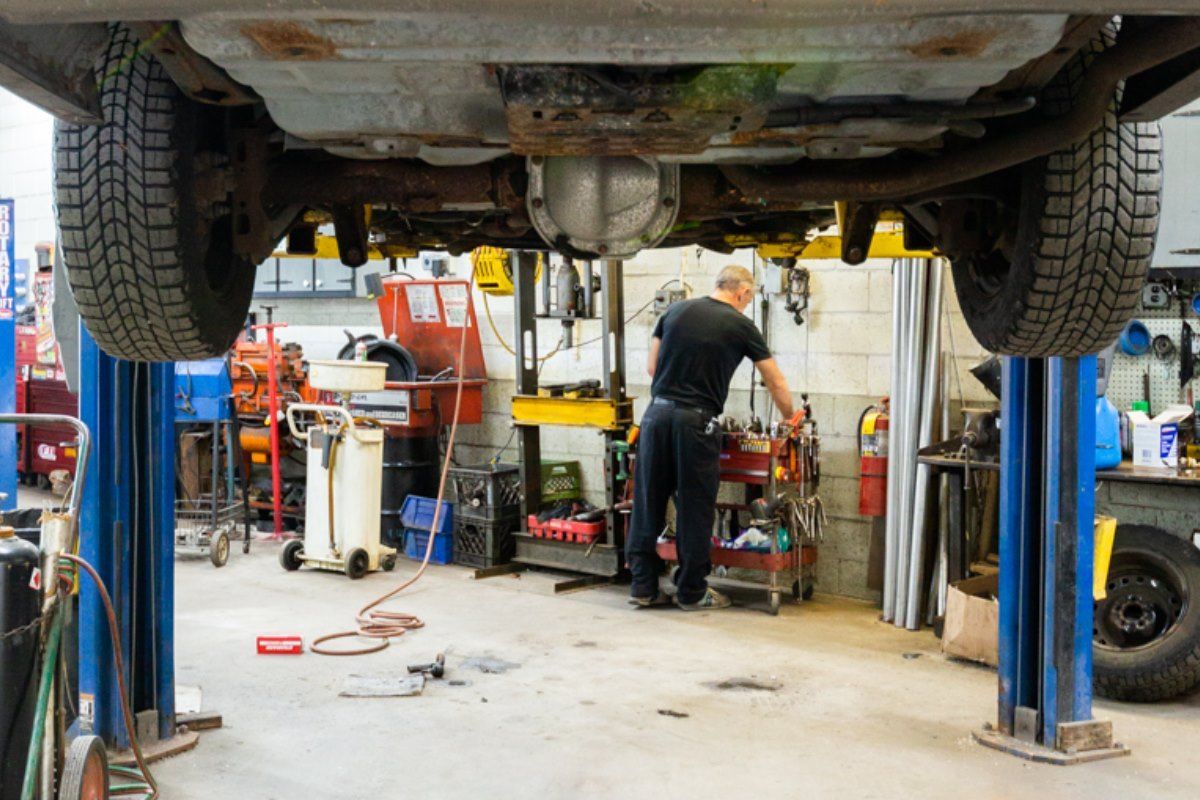 A car raised on a lift in a repair shop; a mechanic works underneath the vehicle.