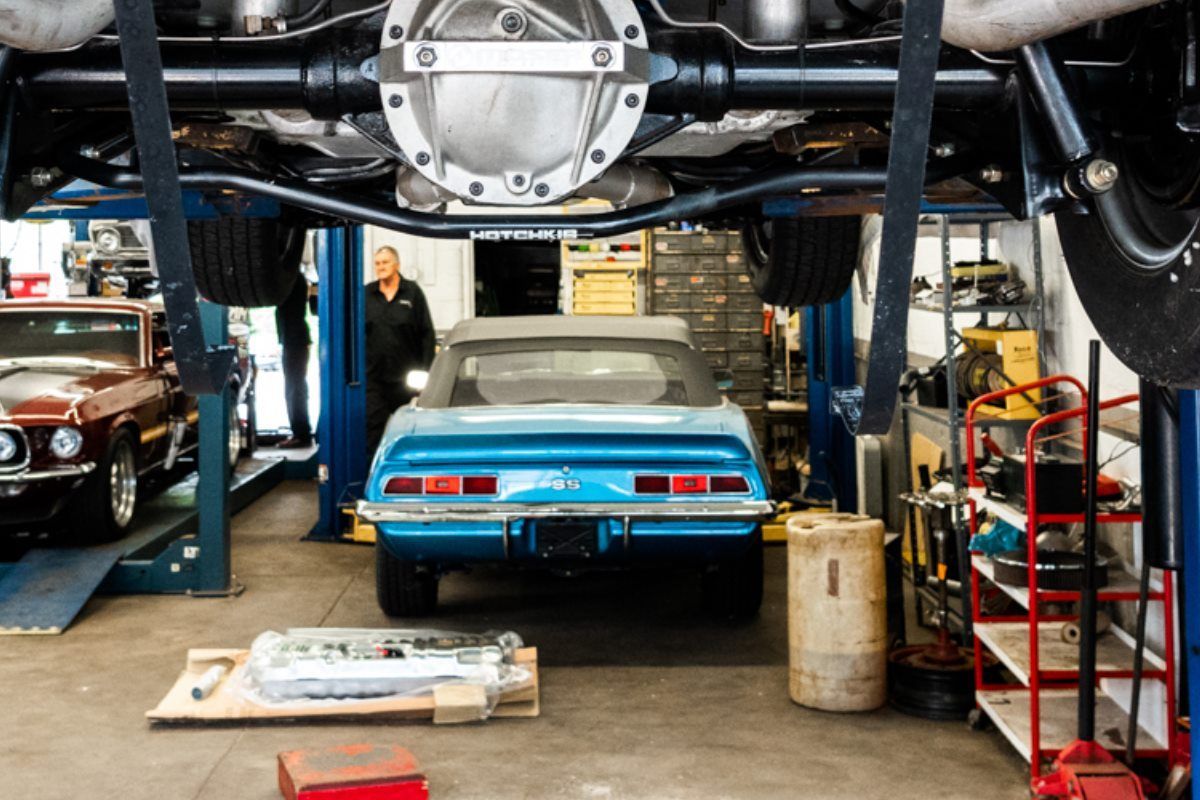 A classic blue car being worked on in a garage, with another car on a lift; a mechanic.