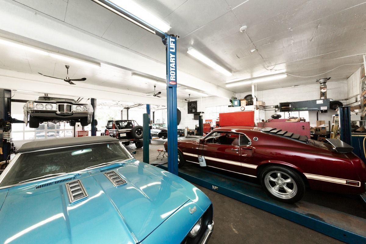 Classic cars in a garage, one blue convertible in foreground, one red on a lift, and another black Jeep on a lift.