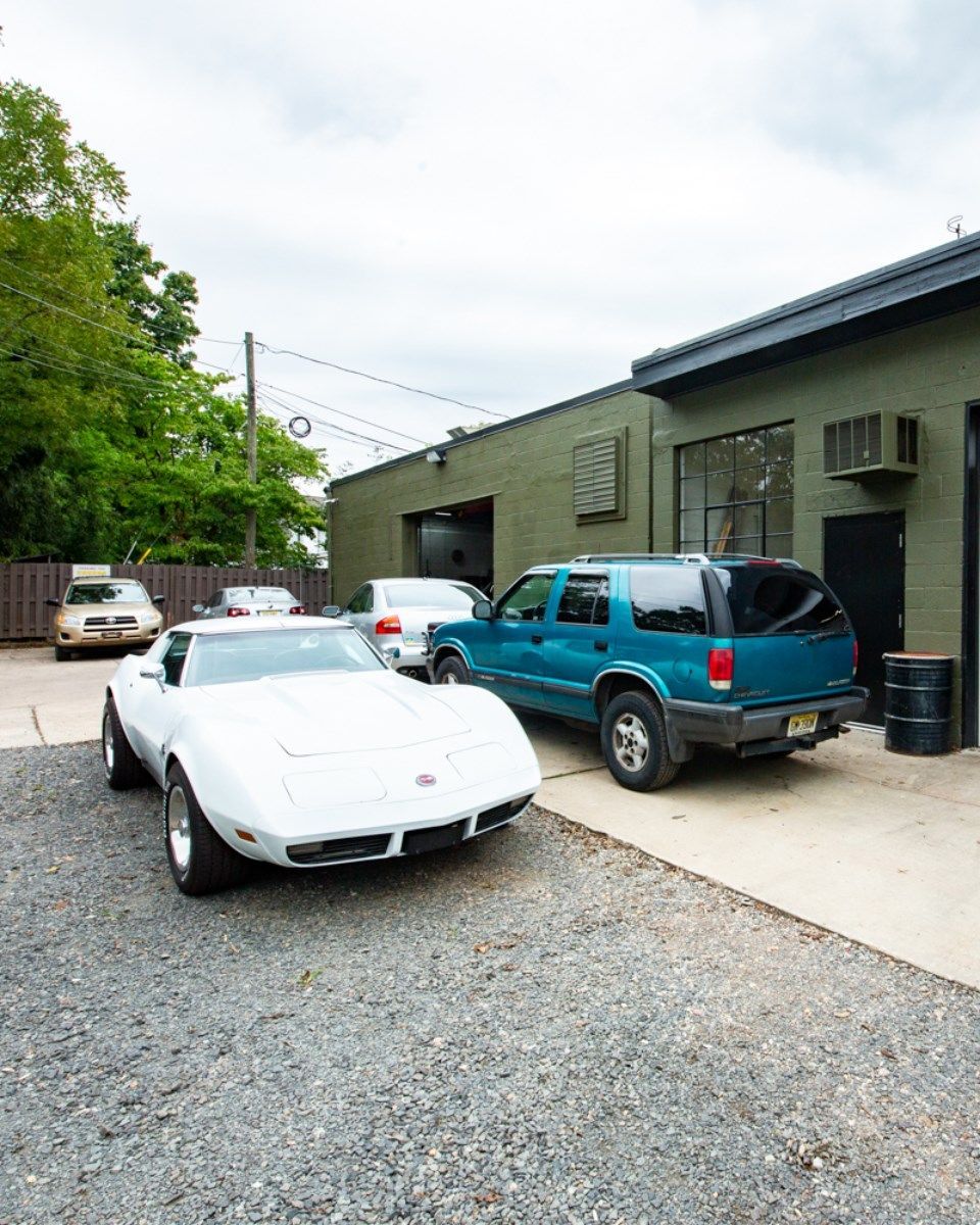 White classic Corvette, teal SUV, and silver car parked in front of a green building with open bay doors.