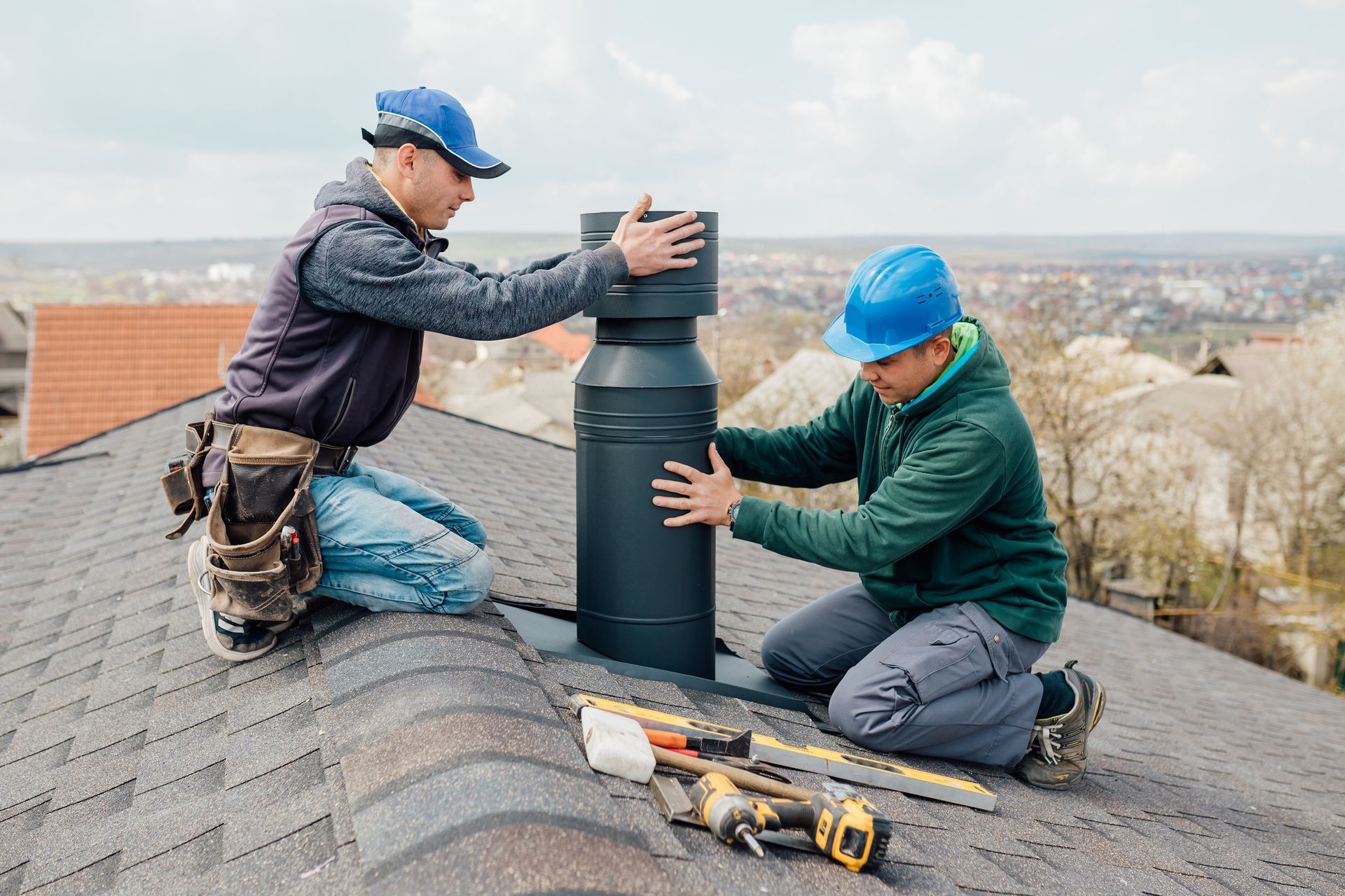 Two workers install a black chimney vent on a roof, with tools nearby.