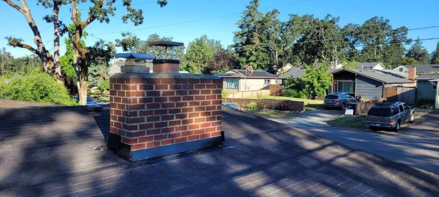 Brick chimney on a shingled roof with houses and trees in the background under a clear blue sky
