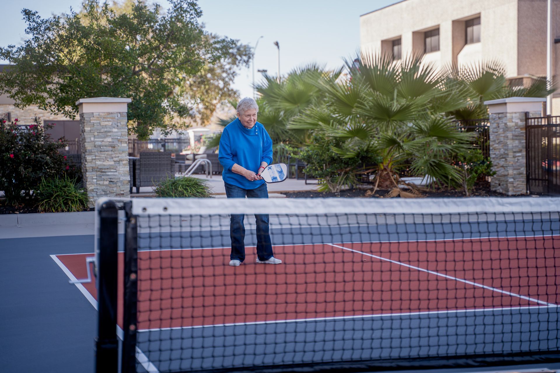 Person playing pickleball on a court with a net. They are holding a paddle, near palm trees.