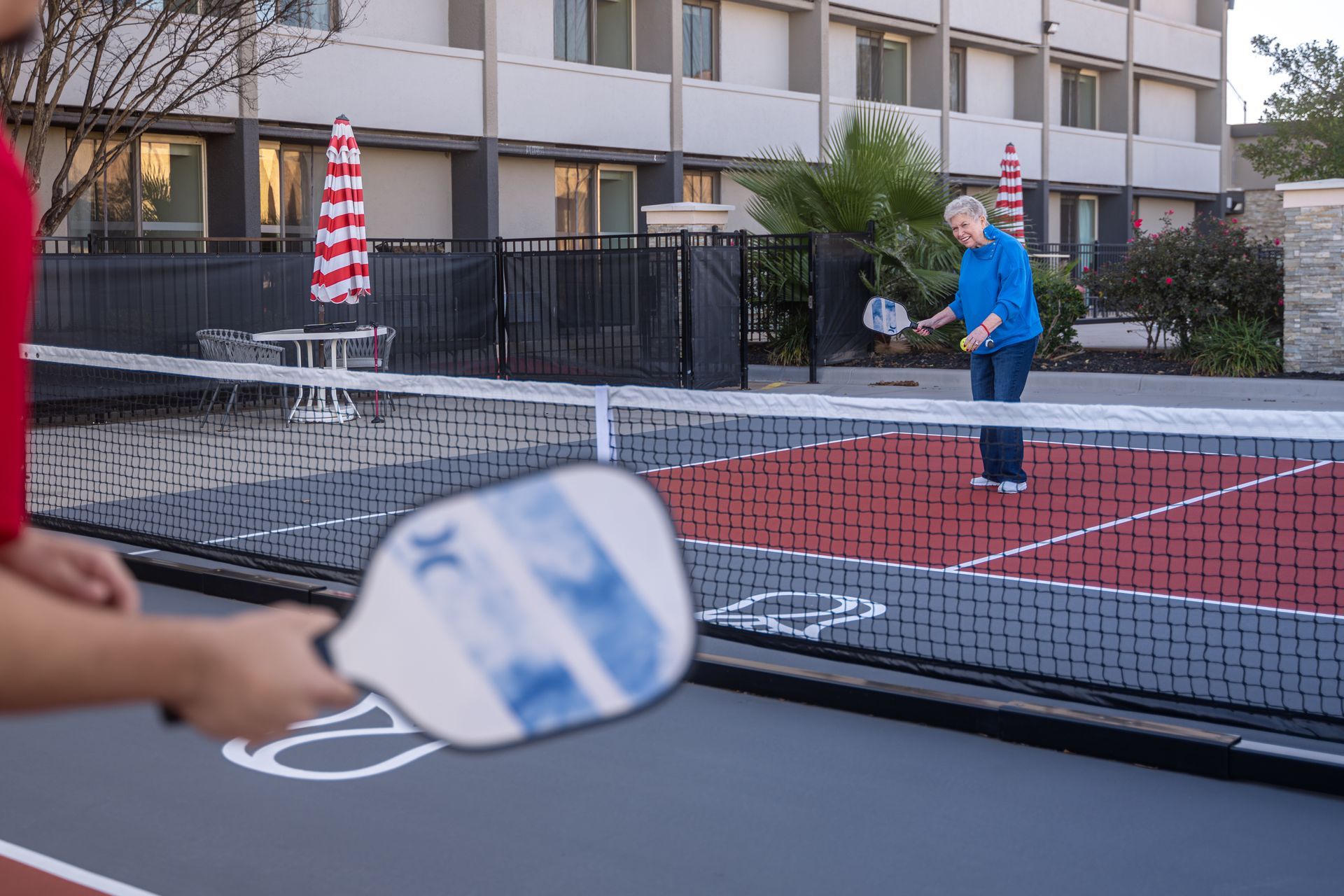 Two people playing pickleball on an outdoor court near a building. One player holds a paddle.