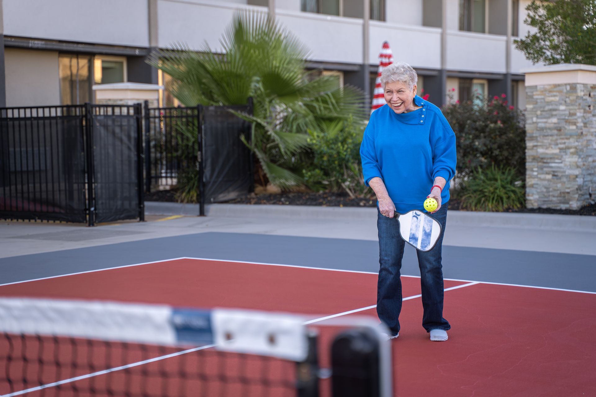 Woman playing pickleball outdoors, smiling. Holding paddle, ball. Red court, net.