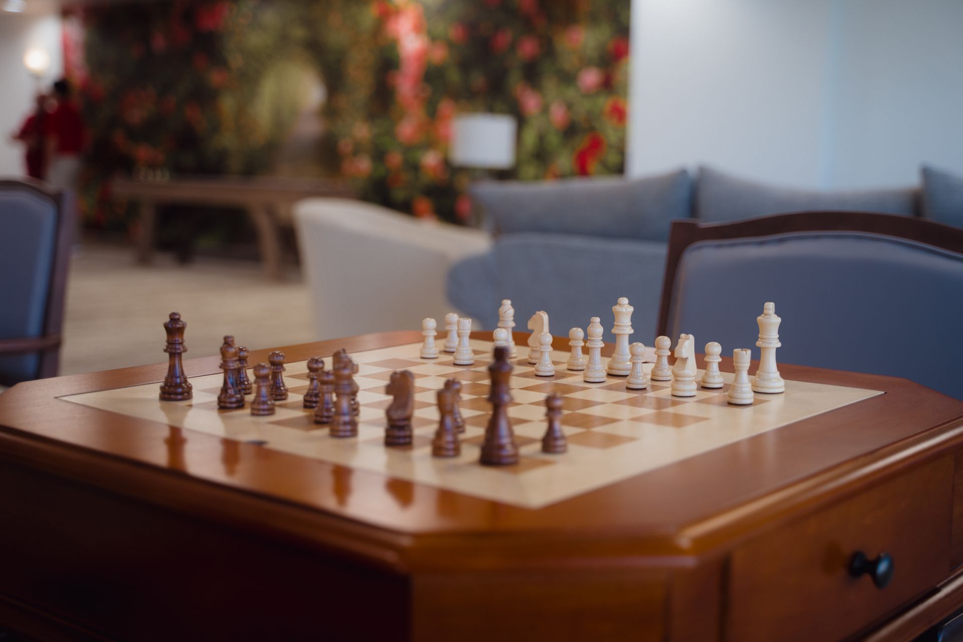 Chessboard on a wooden table, pieces set for a game, in a room with a floral backdrop and a sofa.