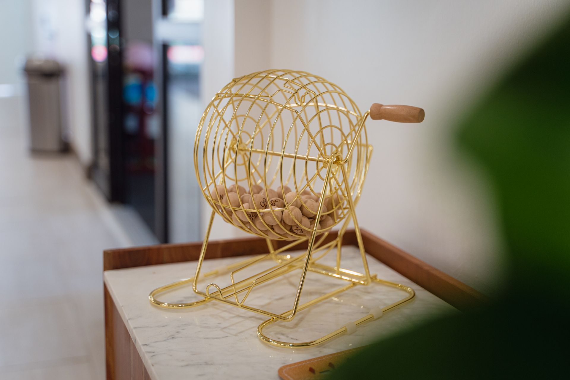 Gold wire raffle drum with wooden handle and beige balls on a marble-topped table.