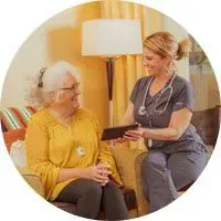 Woman in scrubs consults with an older woman, both looking at a tablet in a home setting.