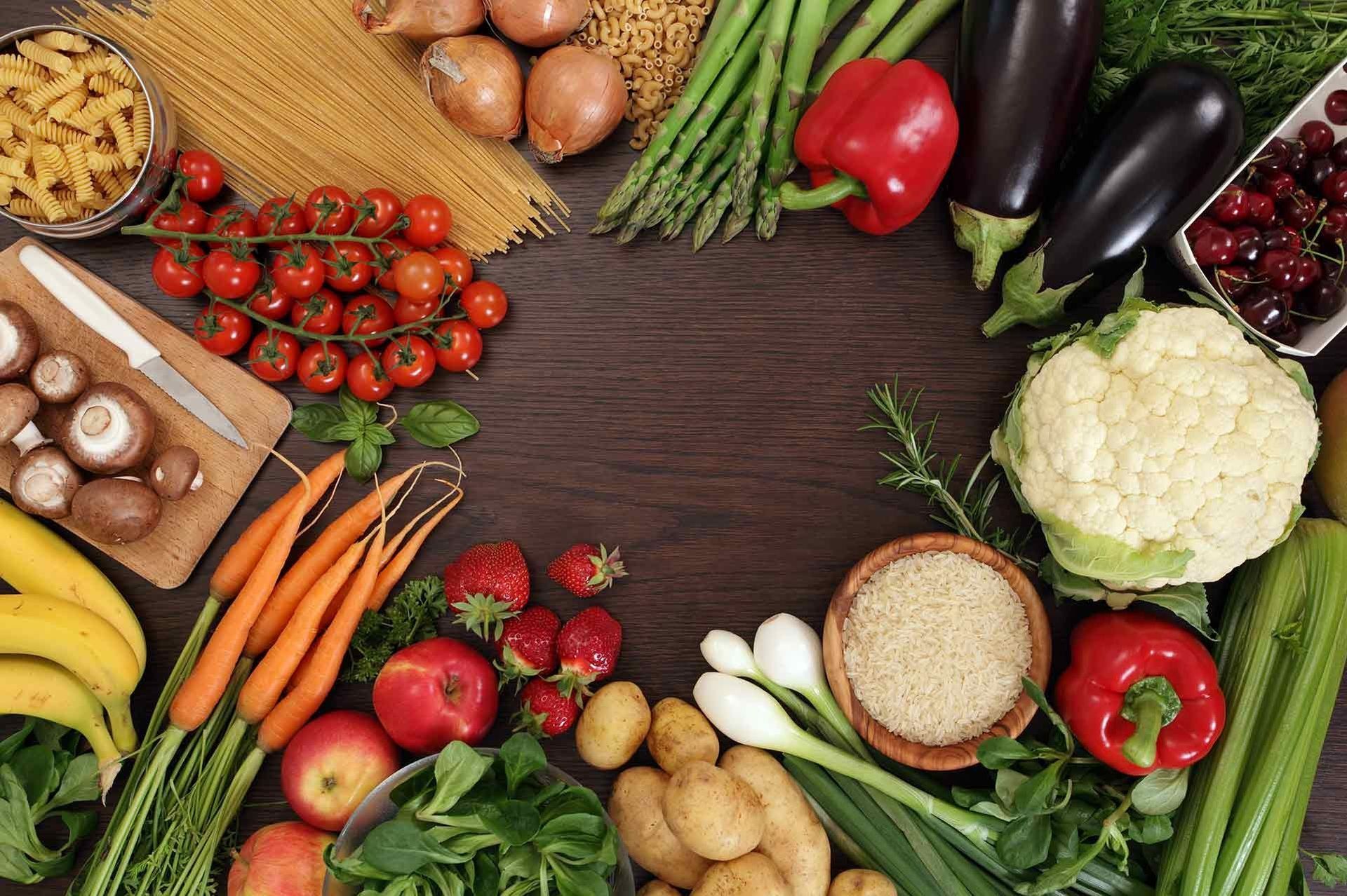 Assortment of colorful fresh produce, including vegetables, fruits, pasta, and grains, arranged on a wooden surface.