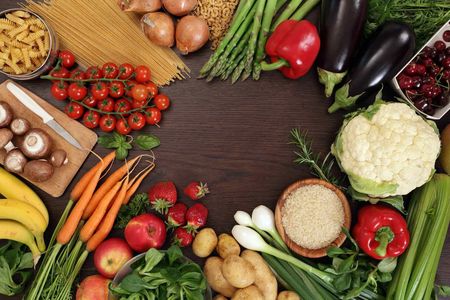 Assortment of colorful fresh produce, including vegetables, fruits, pasta, and grains, arranged on a wooden surface.