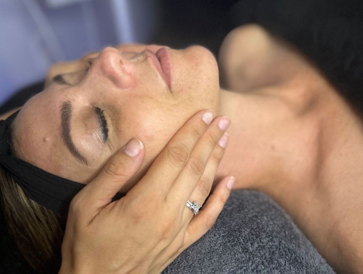 Woman receiving facial treatment; esthetician's gloved hands gently massaging face, eyes closed, in spa setting.