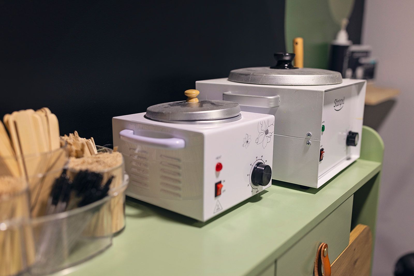 Two white wax warmers on a green counter, with wooden applicators in a clear container.