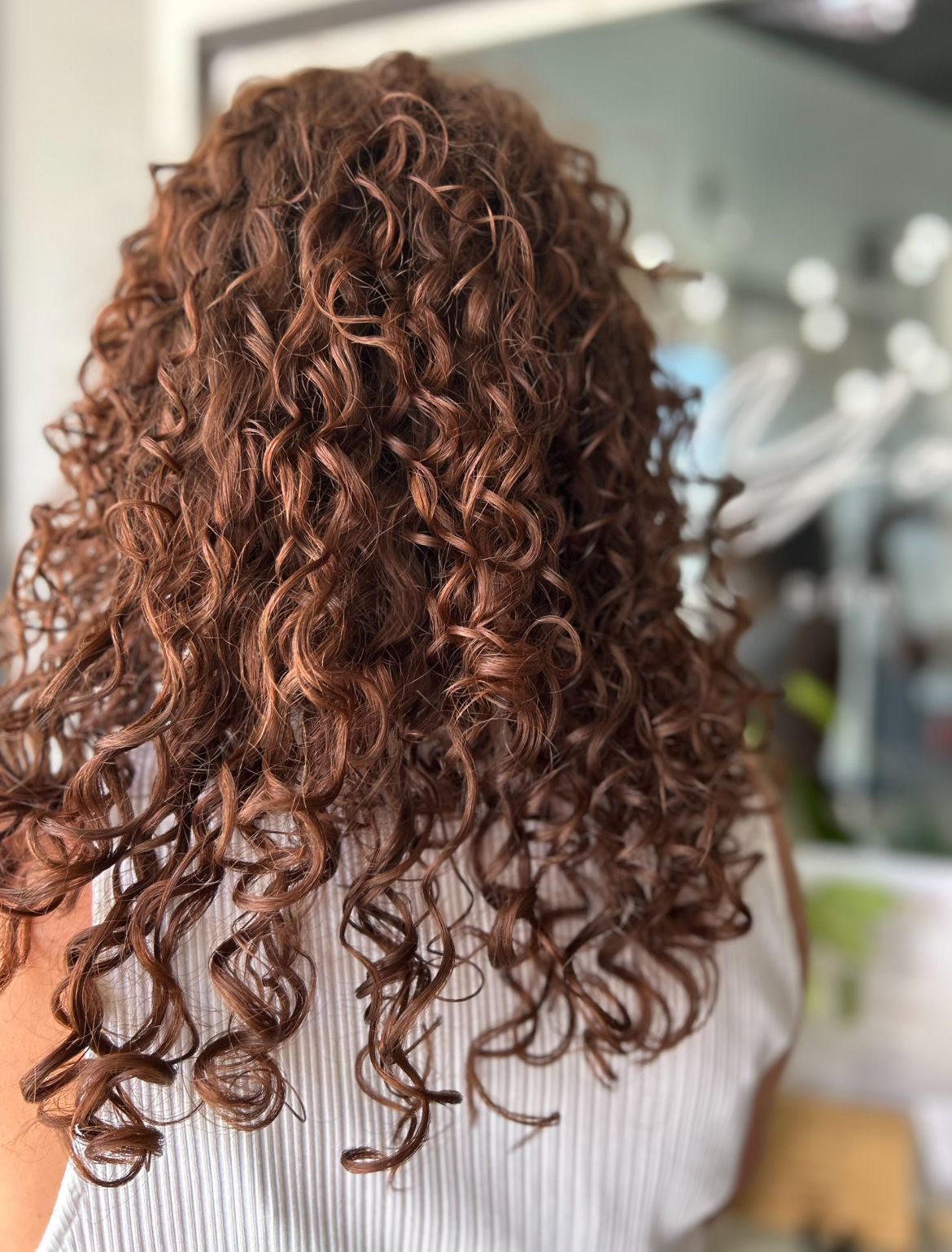 Woman with shoulder-length, reddish-brown, tightly curled hair. Back view, with blurred background.