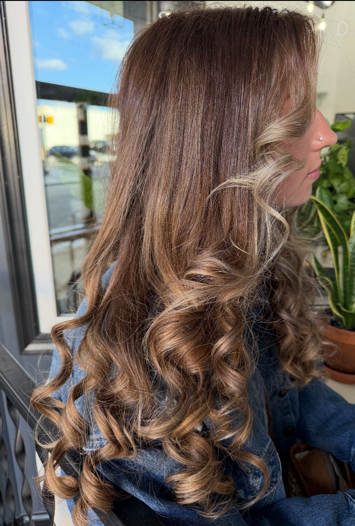 Woman with long brown curly hair, wearing a denim jacket, by a window with a plant.